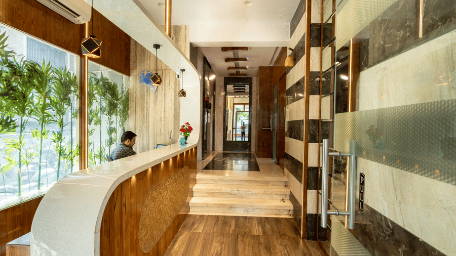 A reception area at Home2 India with a curved front desk, chairs, wall decor, large glass windows, wooden flooring, and a corridor leading inside.