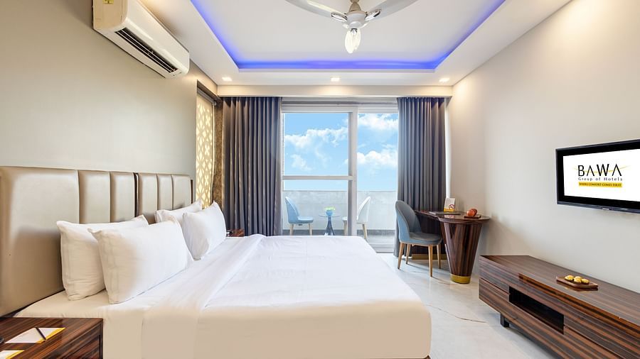 Side view of a king size bed in a room with elegant decor and a balcony view at Hotel Bawa, Gurgaon.