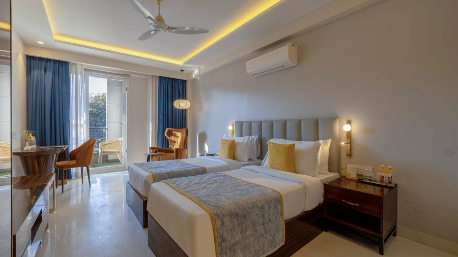 Bright hotel room interior showing twin beds and a glass door leading to a sunlit balcony.