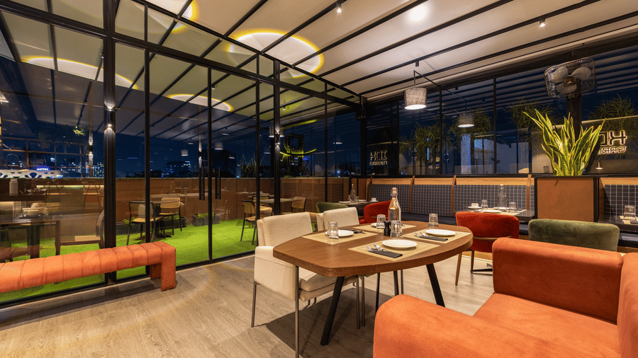 Nighttime view of the dining space at Koi Garden inside Hotel Bawa Gurgaon. A table and chairs sit in the foreground near a glass partition. Plants are placed along the perimeter of the room.