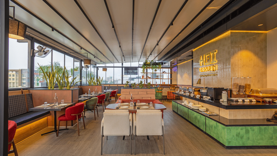 A wide view of the seating layout inside Koi Garden at Hotel Bawa Gurgaon. Dining tables with place settings occupy the central floor area. A serving counter stretches along the right wall under ceiling lights.