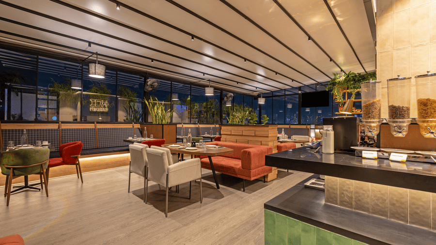 View of the interior layout at Koi Garden restaurant within Hotel Bawa Gurgaon. Tables and seating options span the floor towards a counter with dispensers. Glass panels look out to the exterior surroundings.