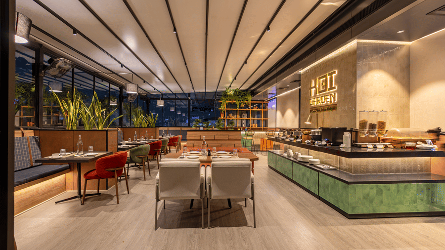 An expansive view of the Koi Garden dining room at Hotel Bawa Gurgaon during the evening. Multiple tables are prepared for guests across the floor. A buffet station is positioned along the right side.