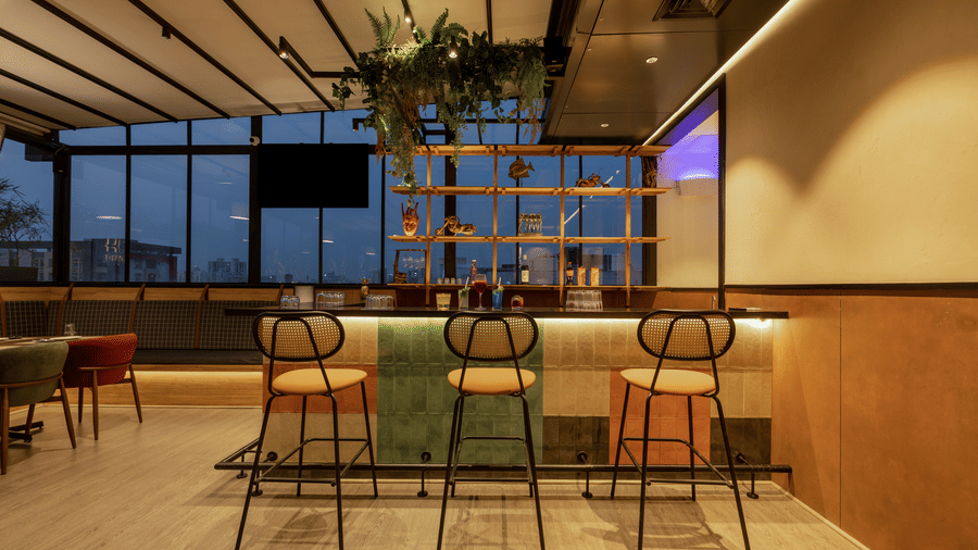 Bar area inside Koi Garden restaurant. Stools are placed along the front of the counter. Shelves with bottles stand beneath the ceiling structure and a hanging plant.