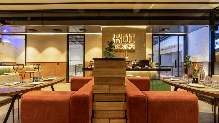 Seating arrangements near the entrance of Koi Garden restaurant at Hotel Bawa Gurgaon. Seating booths face each other, separated by a low divider. The restaurant sign is visible on the wall behind the counter.