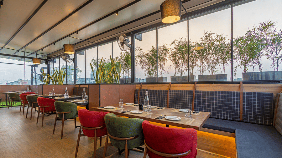 Seating section alongside windows at Koi Garden restaurant. Dining tables with place settings face a row of chairs and a bench. Planters with foliage stand outside the glass partition.