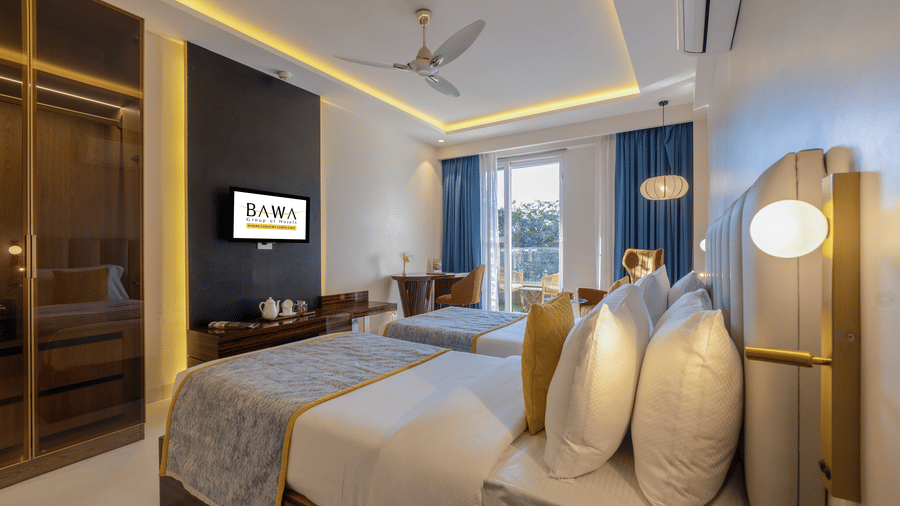 Spacious hotel room featuring two twin beds with gray runners, blue curtains, and warm recessed lighting.