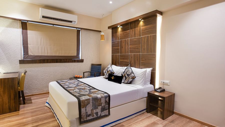 A side corner view of the Deluxe room at Hotel Bawa Regency, Dadar, featuring a plush bed, a window, a small stool beside the bed, and a working desk.