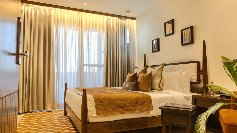 Spacious bedroom with a king size bed dressed in warm toned linen, wooden headboard and soft curtains at Hotel Bawa, Udaipur.