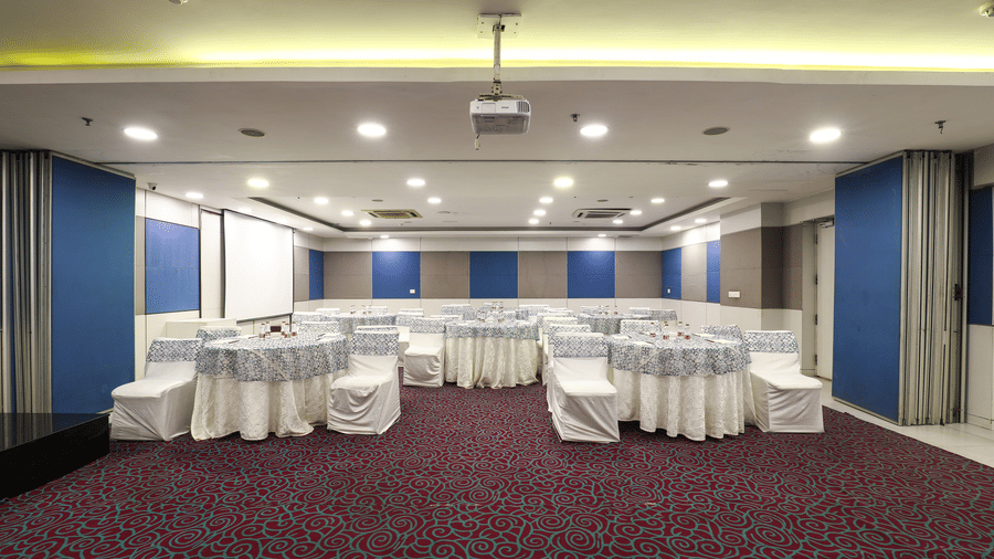 A large and well-lit conference hall with round tables, white chair covers, and a modern projector setup, perfect for professional events and meetings.
