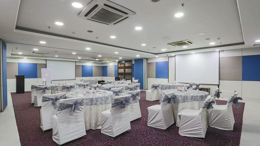 A spacious and elegantly designed conference hall with round tables and chairs draped in white covers, featuring advanced lighting and projection equipment.