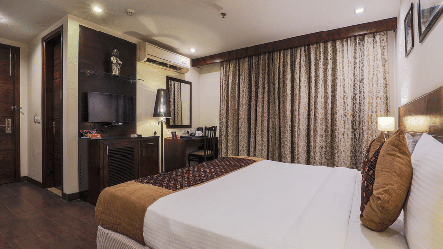 A well-lit room at Hotel Bluestone Prime (Nehru Place), New Delhi featuring a large white bed, a desk area, and a wall of patterned curtains covering the windows.