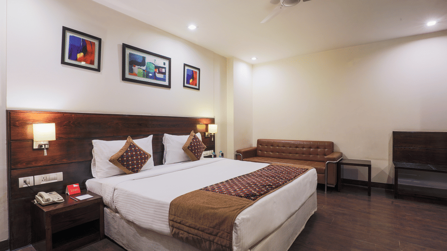 A spacious room at Hotel Bluestone Prime (Nehru Place), New Delhi, featuring a double bed with a brown runner, artistic frames placed above the bed and a small, dark sofa arranged in the corner.