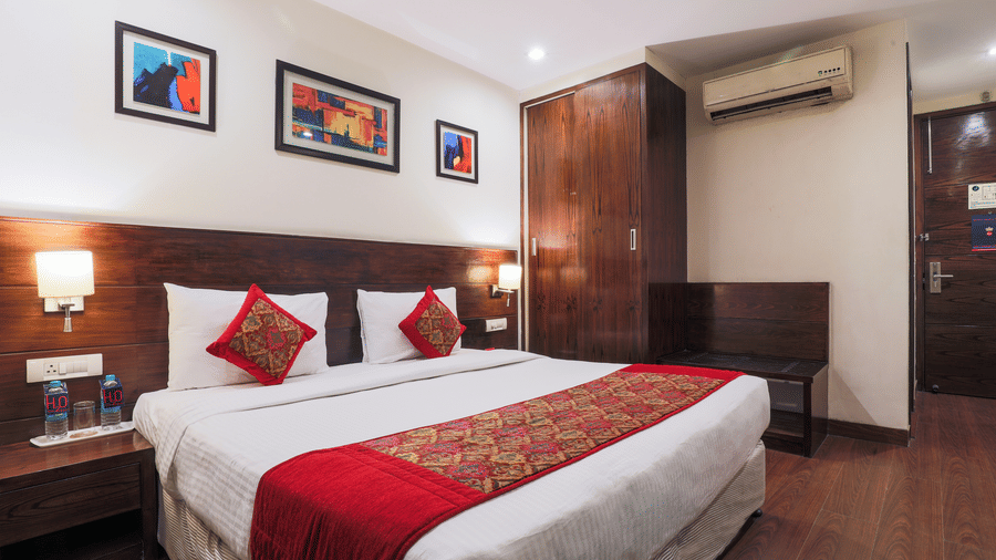A neatly arranged hotel room with a double bed displaying a bright red runner, a small dark wooden wardrobe and artistic frames placed above the bed - Hotel Bluestone Prime (Nehru Place), New Delhi