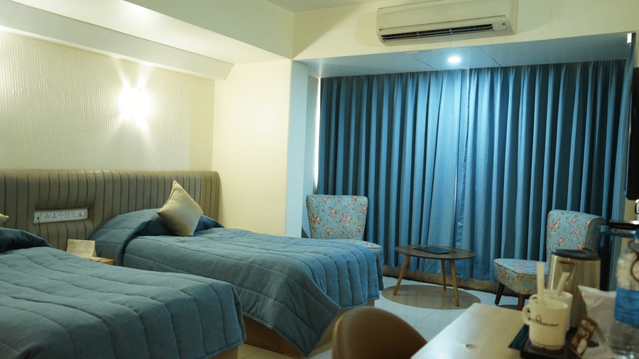A room with twin beds, spreads, pillows, chairs, and a large window covered with curtains.