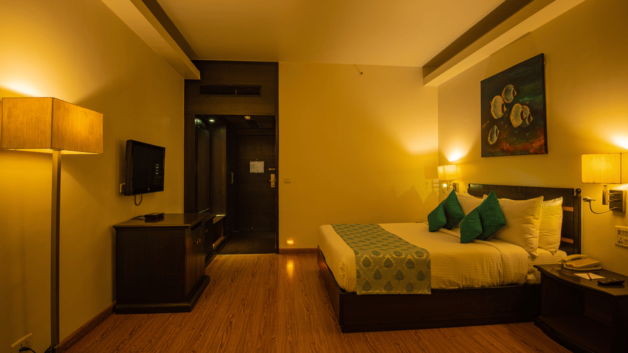 Hotel room at Hotel Kanha Shyam with a bed dressed in white linen, a wall-mounted artwork, a wall-mounted television and warm ambient lighting.