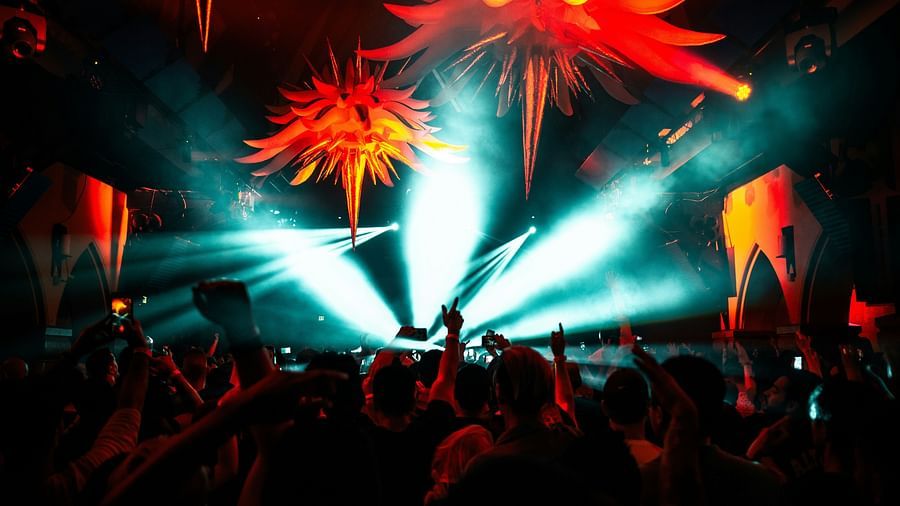 A vibrant nightclub scene with a crowd of people dancing under large, spiky, orange and red decorative lights. Bright beams of light cut through the smoky atmosphere, creating an intense, energetic vibe.