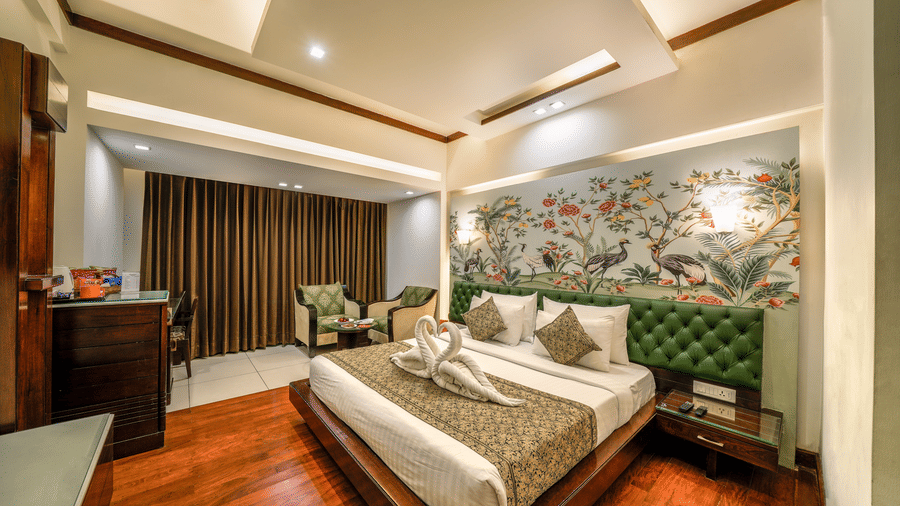 A well-lit, Hotel Saffron Leaf, Dehradun's room featuring a large bed with a runner and matching pillows against a decorative patterned wallpaper.