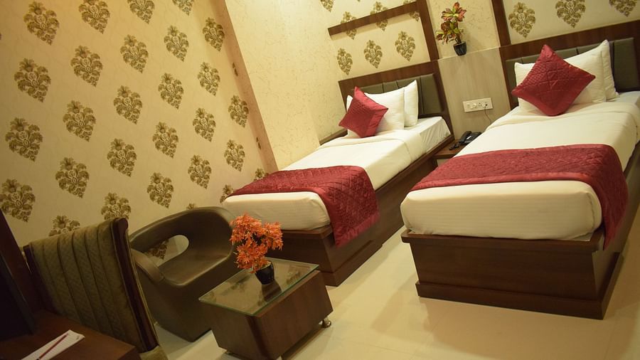 A spacious twin-bed room with patterned wallpaper, a glass table with a cushioned chair, and a dining setup at Hotel Uday Palace.