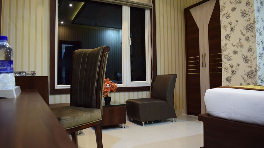 A spacious room with a wooden table and chair, large windows with pulled-up curtains, and a cushioned chair with a mini table in front at Hotel Uday Palace.