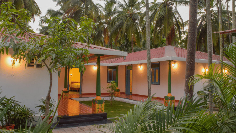 A white resort cottage with a red roof and blue window frames, surrounded by palm trees and green lawns - Ibex Resorts, Coimbatore (Kakarla)