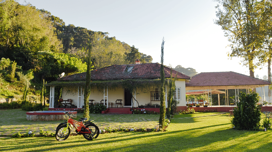 A large white house with a red roof, surrounded by an expansive green lawn and tall trees, with a red bicycle parked on the grass and hills in the background under a bright sky.