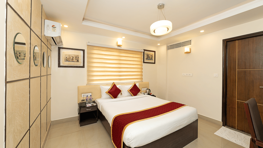 The Deluxe Room of Icon Regency by Bhagini, Whitefield showcasing a double bed with crisp linens, wardrobe, an air conditioning, ceiling lights, closed wooden door, and tiled flooring.