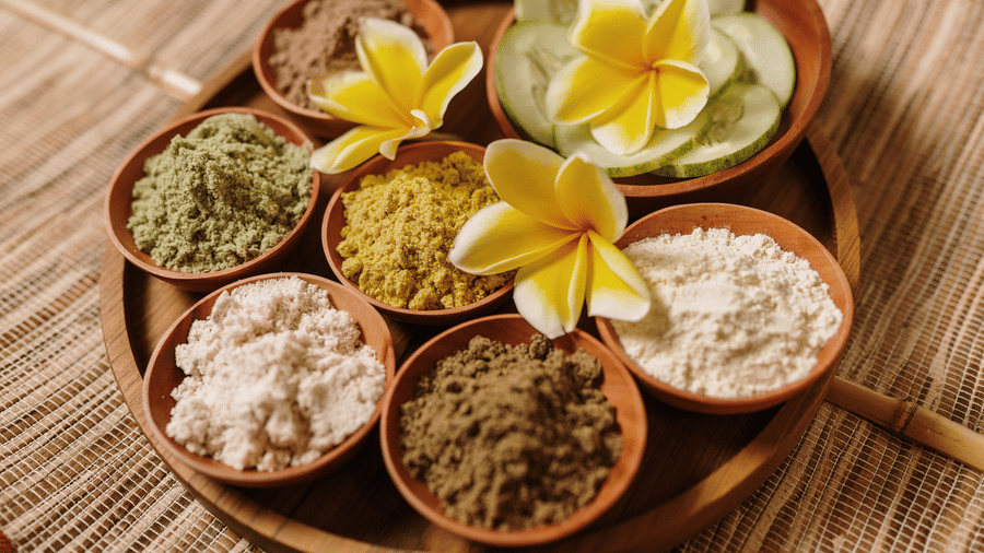 Assorted natural body scrubs and spa ingredients arranged in wooden bowls with tropical flowers.
