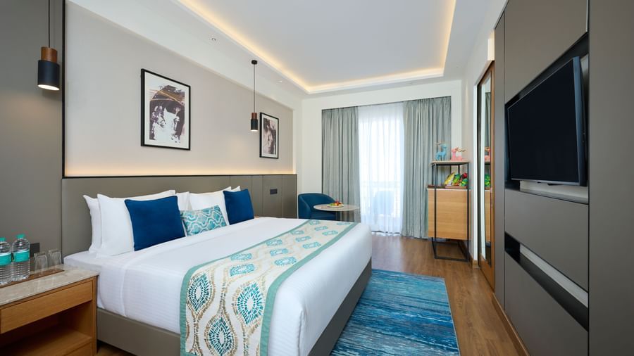 A stylish hotel bedroom features a king-sized bed with blue and patterned cushions and a throw, a blue rug, a wall-mounted TV, a mini-fridge unit, and a chair by the window. - Iris Sarovar Portico, Sector 85, Gurugram