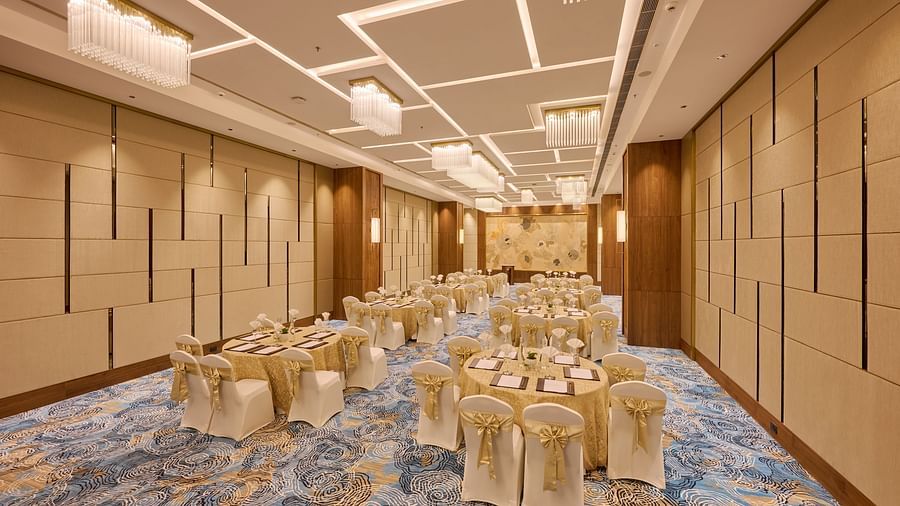 A well-lit banquet hall features multiple round tables, each elegantly set with tableware and surrounded by chairs draped with gold fabric, ready for a formal gathering. - Iris Sarovar Portico, Sector 85, Gurugram