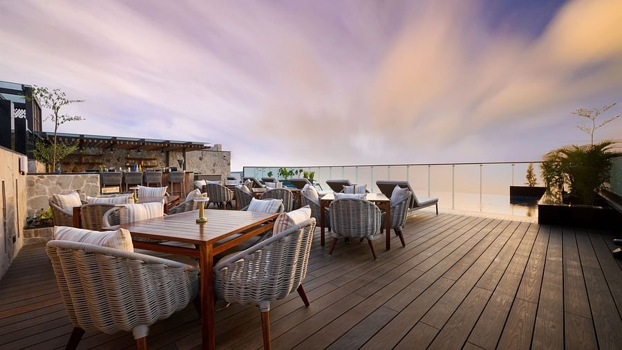 A rooftop terrace with wooden decking features several dining tables and comfortable chairs with white cushions, offering an open-air space under a partly cloudy sky. - Iris Sarovar Portico, Sector 85, Gurugram