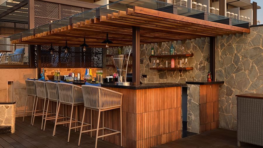 A stylish outdoor bar area with a wooden countertop and stools is set up, featuring shelves stocked with bottles and glasses, ready to serve drinks in a relaxed setting. - Iris Sarovar Portico, Sector 85, Gurugram