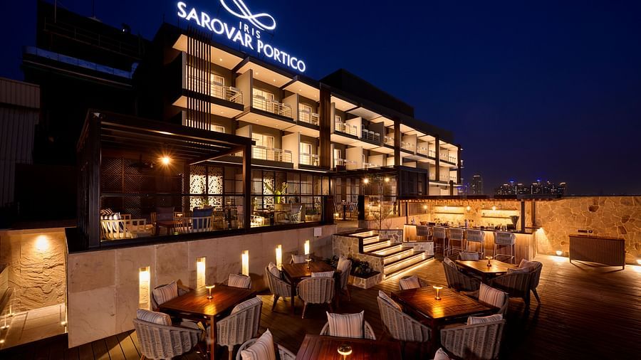 A stylish rooftop bar and dining area is illuminated at night, with tables and chairs arranged on a terrace overlooking the cityscape, with the hotel's signage visible above. - Iris Sarovar Portico, Sector 85, Gurugram