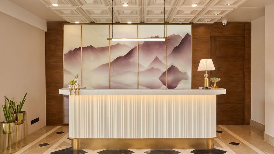 A stylish hotel reception desk with a white countertop and gold trim is positioned in front of a decorative wall panel featuring an abstract mountain scene. - Iris Sarovar Portico, Sector 85, Gurugram