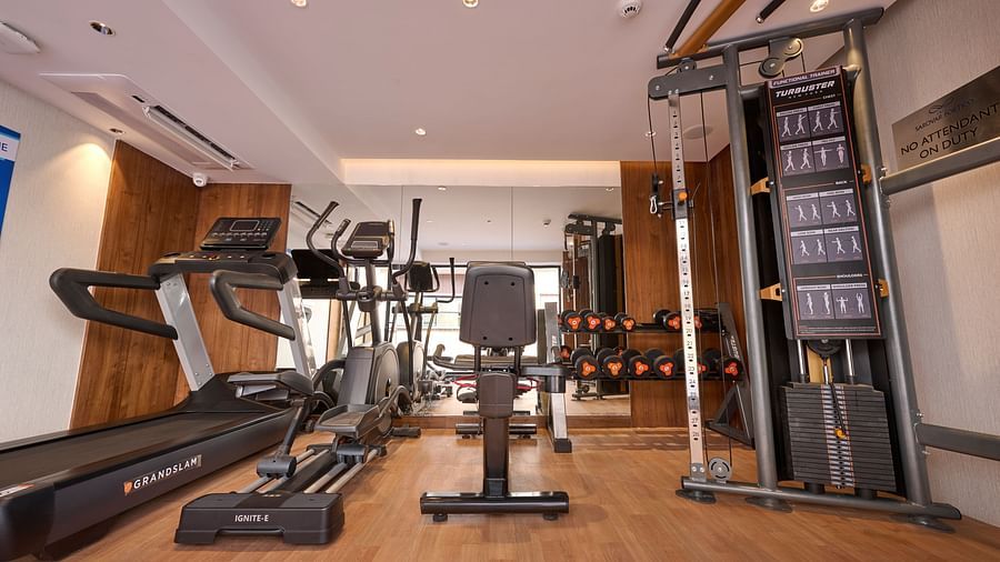 A well-equipped hotel gym features a treadmill, elliptical machine, weight bench, and a cable machine with various weight plates, all on a wooden floor. - Iris Sarovar Portico, Sector 85, Gurugram