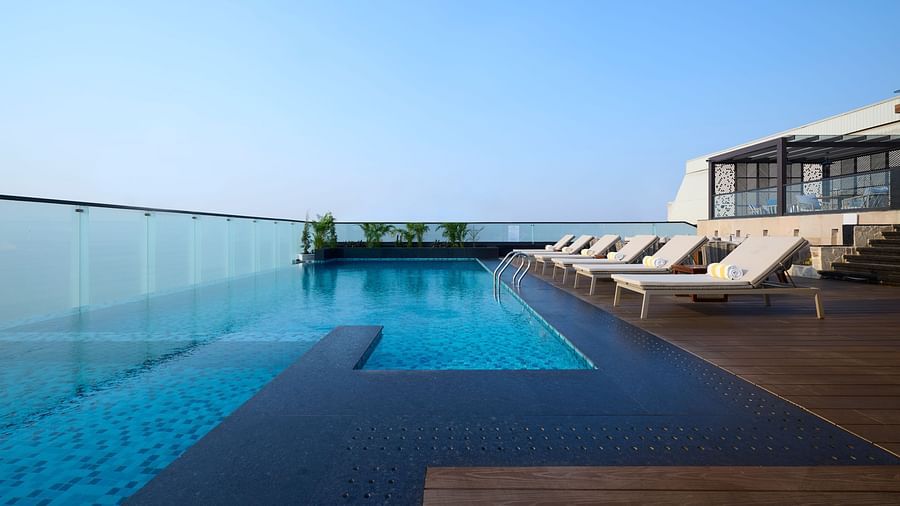 A rooftop infinity pool with clear blue water extends to the edge of the building, offering a view of the clear sky, with lounge chairs lined up on the decking. - Iris Sarovar Portico, Sector 85, Gurugram