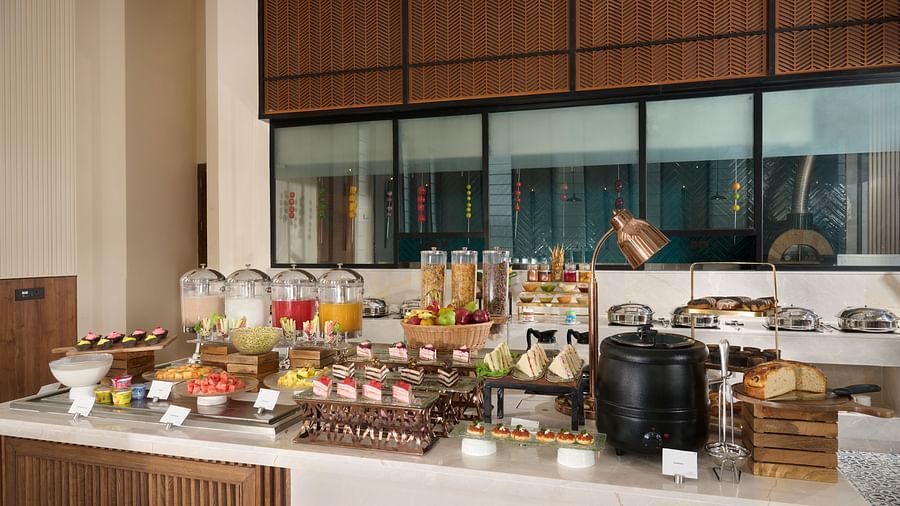 A breakfast buffet is laid out on a long white counter, offering a selection of cereals, pastries, fruit, and juices, ready for hotel guests. - Iris Sarovar Portico, Sector 85, Gurugram