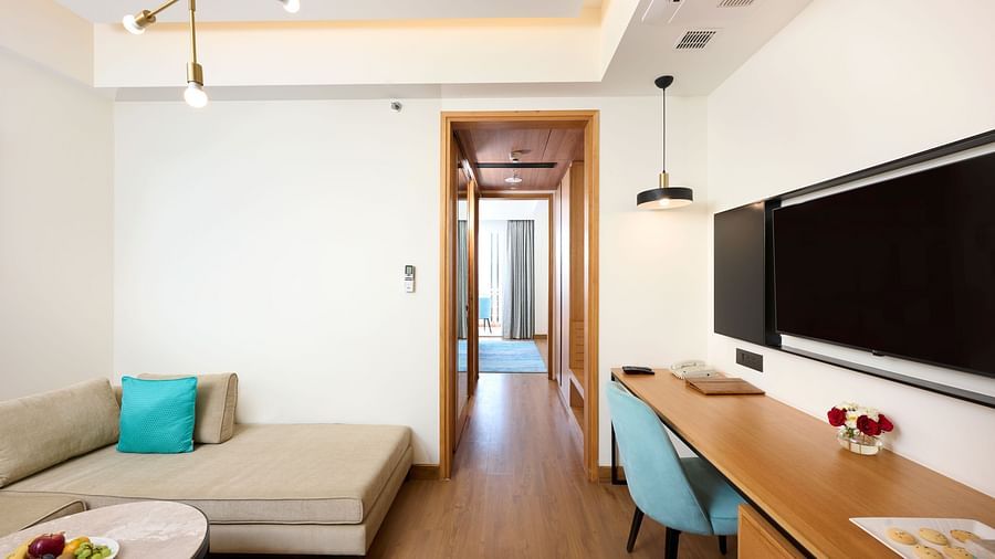 A comfortable hotel room features a sofa with a turquoise cushion, a desk with a blue chair, a wall-mounted television, and a doorway leading to another area. - Iris Sarovar Portico, Sector 85, Gurugram