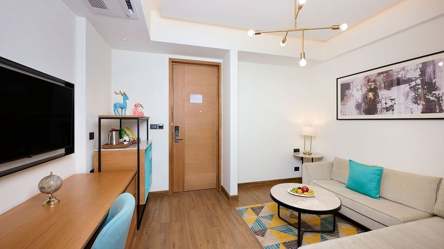 A comfortable hotel suite includes a sofa with a turquoise cushion, a desk with a blue chair, a wall-mounted TV, and a round coffee table on a colourful geometric rug. - Iris Sarovar Portico, Sector 85, Gurugram