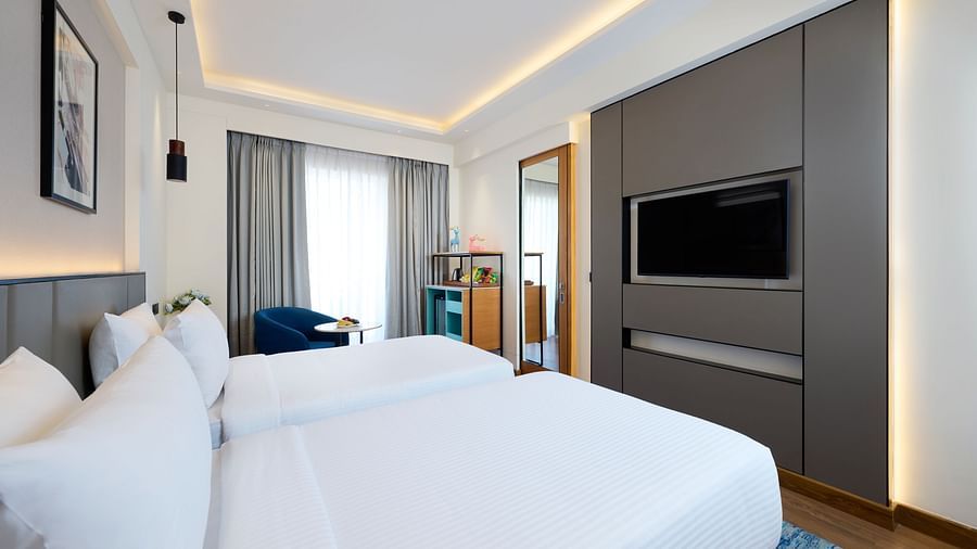 A bright hotel room with two single beds includes a wall-mounted television, a mini-fridge unit, a chair by the window with grey curtains, and framed artwork on the wall. - Iris Sarovar Portico, Sector 85, Gurugram