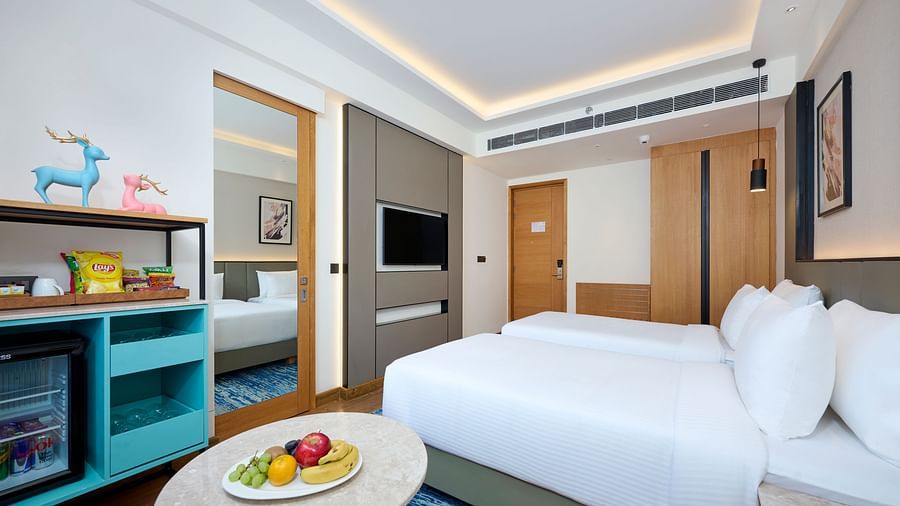 A hotel room with two single beds features a mini-fridge unit, a round table with fruit, a wall-mounted TV, and a glimpse into an adjacent room through an open doorway. - Iris Sarovar Portico, Sector 85, Gurugram