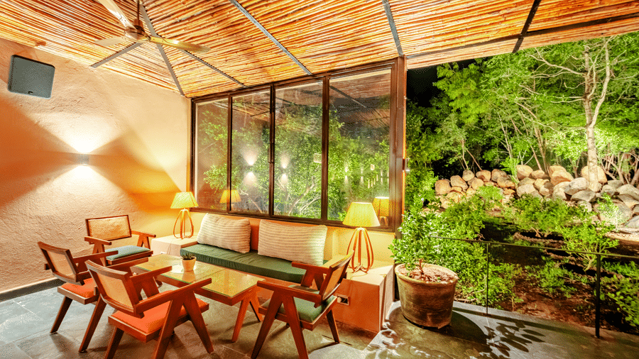 An image showing wooden chairs and table placed aesthetically inside a room with bamboo roofing, where the garden view is at an enjoyable distance