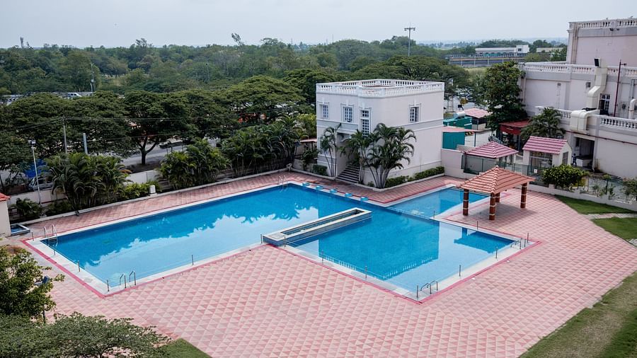 A large outdoor swimming pool with clean water, surrounded by a tiled deck, nearby buildings, and trees under a cloudy sky at Jenneys Residency