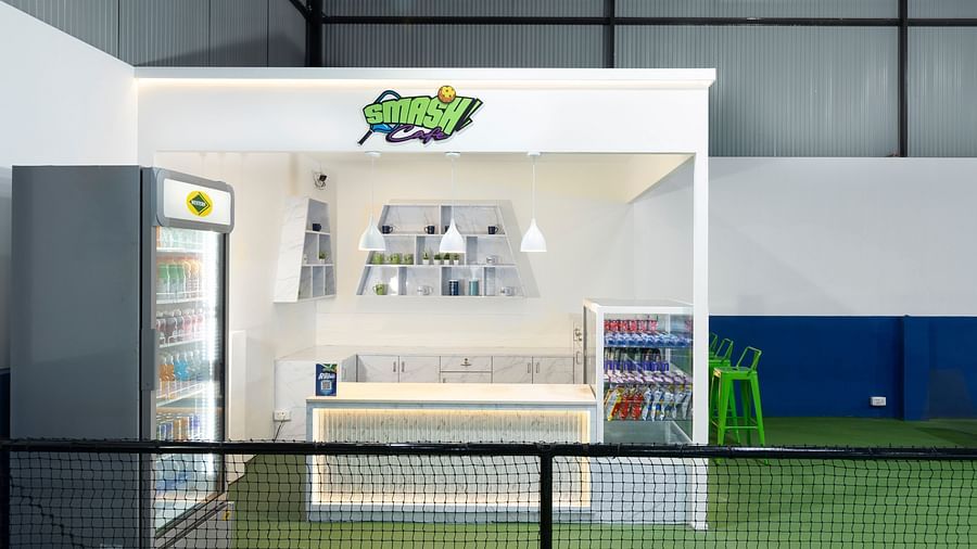 Modern refreshment counter labelled 'Smash Café' with a fridge, seating area, and shelves, located inside the indoor sports facility at Jenneys Residency, Coimbatore.