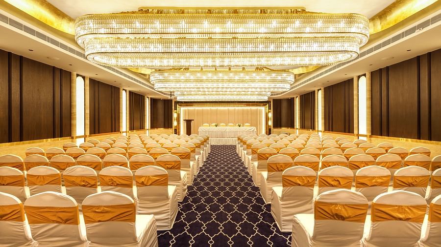 Elegant banquet hall with rows of white and gold-covered chairs facing a stage, illuminated by chandeliers, at Jenneys Residency, Coimbatore.
