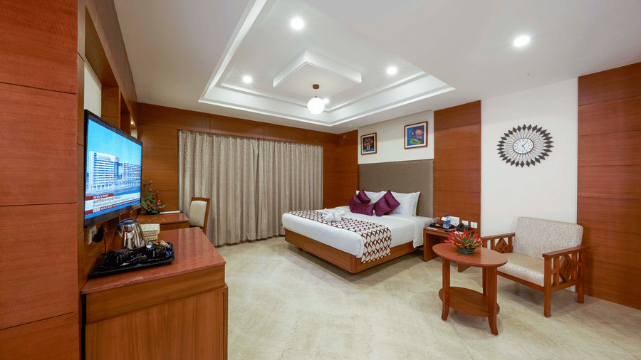 A suite at JP Hotel Chennai featuring a plush bed next to a seating space facing the TV over a wooden counter, and a window by the bed.