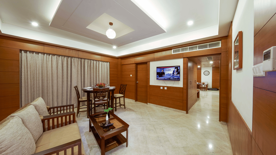 JP Suite at JP Hotel Chennai featuring living area with wooden furnishings, sofas with coffee table facing the TV on the wall next to which is a 4-seater dining table