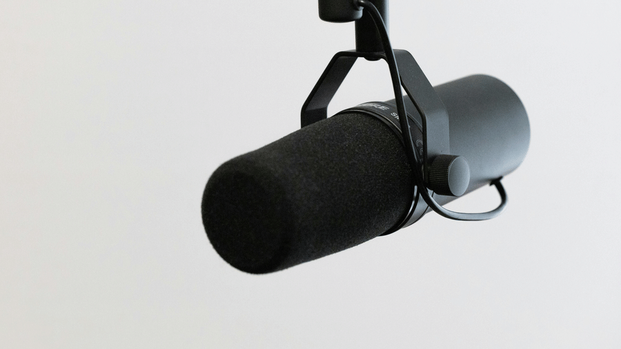 A professional black studio microphone suspended with a stand against a plain light-coloured background.