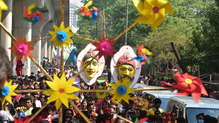 A procession on a road with DIY fans in the foreground and people carrying busts on a pole next to parked cars during Poila Boishakh.