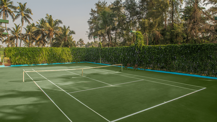 Green outdoor tennis court lined with hedge fencing and tall trees at Kenilworth Resort & Spa, Goa.